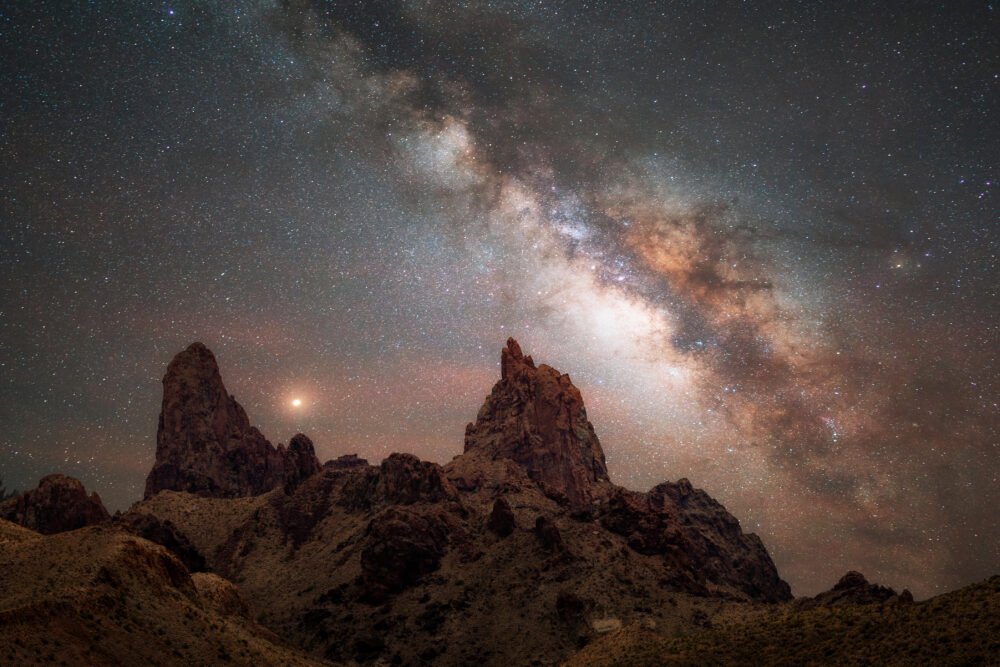 A stunning photo of the Milky Way galaxy over a desert landscape, with two rock formations resembling mule ears in the foreground. The night sky is filled with stars and nebulae, creating a contrast with the dark silhouette of the rocks and plants. The photo showcases the beauty and mystery of the cosmos, as well as the skill and creativity of Andy Morgan Photography.