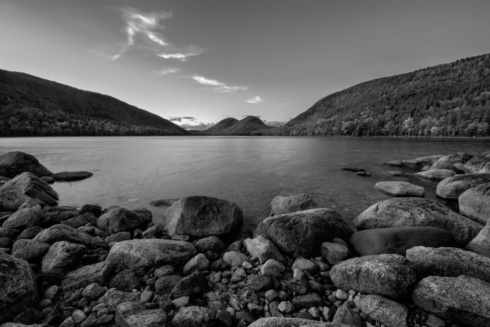 Black and white photo of Jordan Pond in Acadia National Park. Rocky show leads your eyes across the pond to two small rounded mountain peaks in the distance.