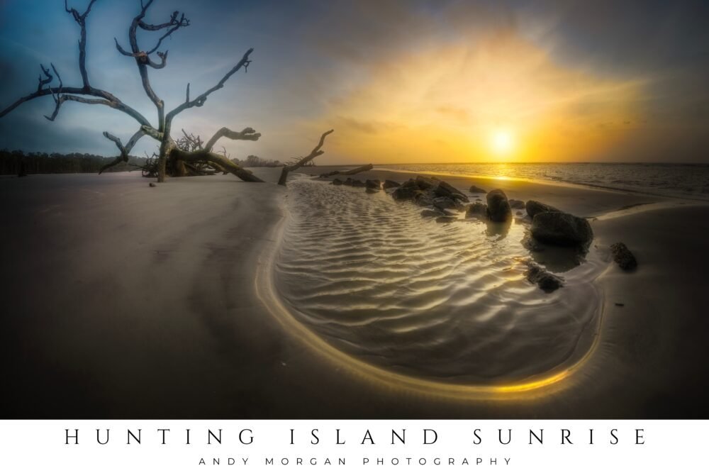 A wide beach scene at sunrise with a dramatic sky of orange and blue, featuring large pieces of dark driftwood on the wet sand at Hunting Island State Park, South Carolina.