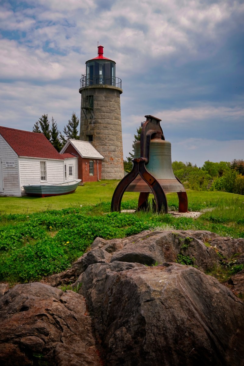 Monhegan Island Lighthouse, its associated keeper's house, and a large metal fog bell on a grassy hill under a partly cloudy sky