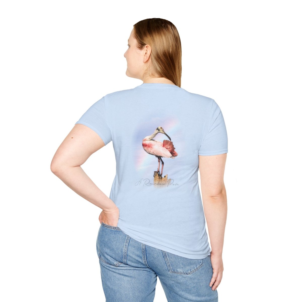 Back view of a person wearing a light blue t-shirt featuring a graphic of a pink roseate spoonbill bird standing on a wooden post, with a subtle rainbow arching behind it. The person has their left hand in their back pocket and is looking over their right shoulder.