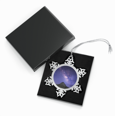 A snowflake-shaped pewter ornament featuring a photograph of the Milky Way galaxy arching above Mitre Peak under a star-filled night sky, displayed in an open black gift box.