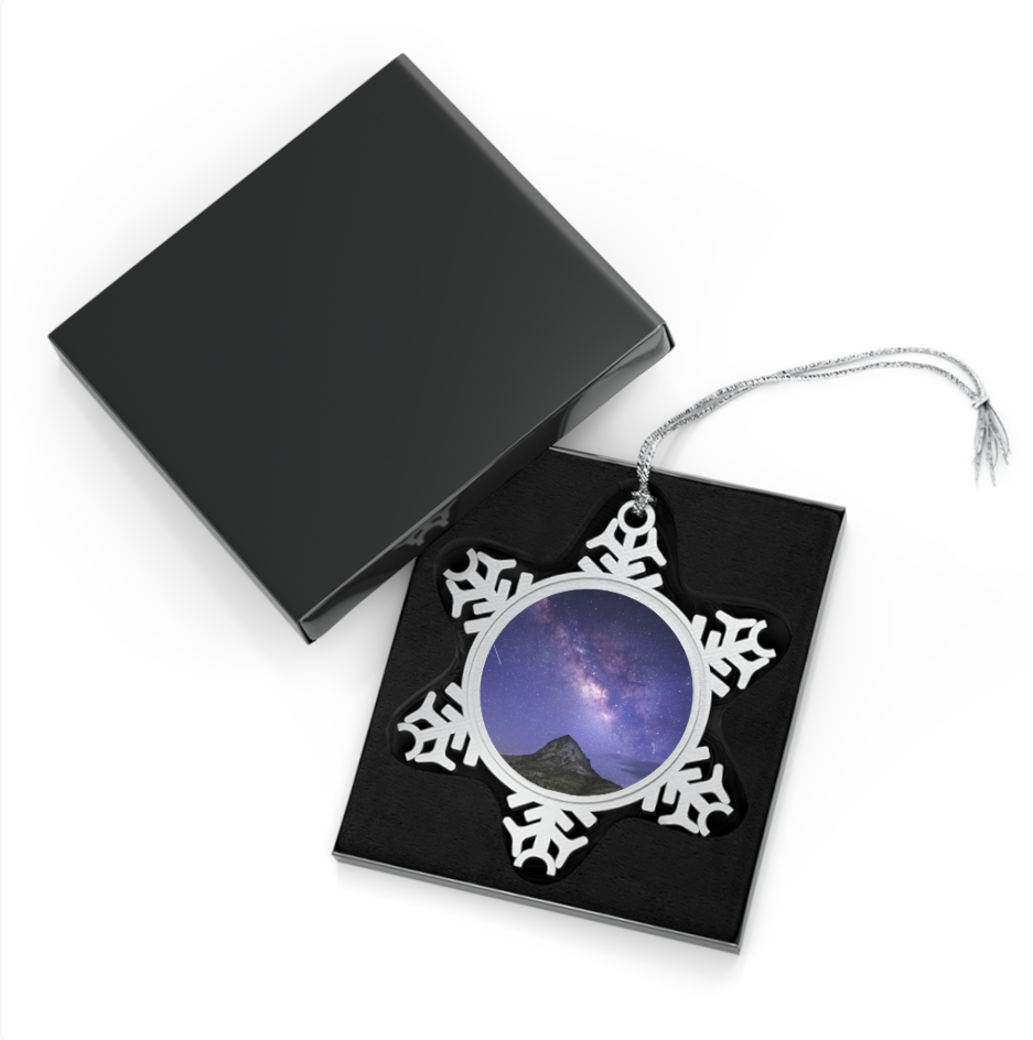 A snowflake-shaped pewter ornament featuring a photograph of the Milky Way galaxy arching above Mitre Peak under a star-filled night sky, displayed in an open black gift box.