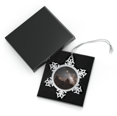 A snowflake-shaped pewter ornament featuring a vibrant photograph of the Milky Way galaxy stretching across a dark night sky above the distinct rock formations of Mule Ears Peaks, with warm light on the horizon.
