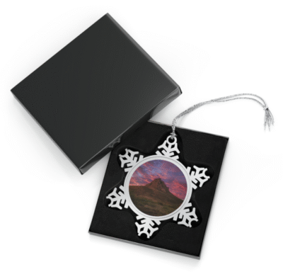 A snowflake-shaped pewter ornament featuring a photograph of Mitre Peak under a vibrant, fiery sunrise with pink and red clouds.