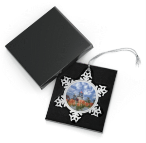 A snowflake-shaped pewter ornament featuring a photograph of the Monhegan Island Lighthouse with a red roof, surrounded by a foreground of colorful tulips, under a sky filled with fluffy white clouds.