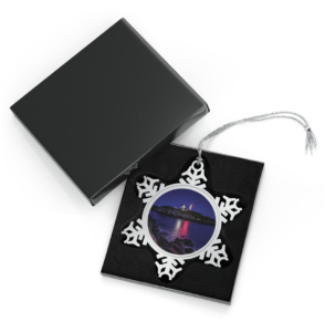 A snowflake-shaped pewter ornament featuring a vibrant photograph of Cape Neddick "Nubble" Lighthouse illuminated at dusk, with waves crashing on the rocks and a colorful sky.