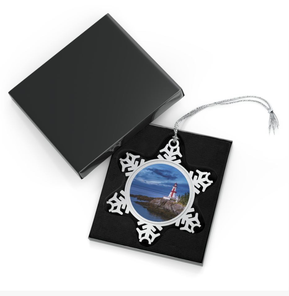 A snowflake-shaped pewter ornament featuring a photograph of the red and white striped Head Harbour Lighthouse (East Quoddy Head Lighthouse) on a rocky coast at dusk, with dramatic blue clouds.