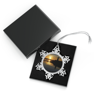 A snowflake-shaped pewter ornament featuring a photograph of the white Mulholland Point Lighthouse silhouetted against a vibrant sunrise, reflected in the calm water.