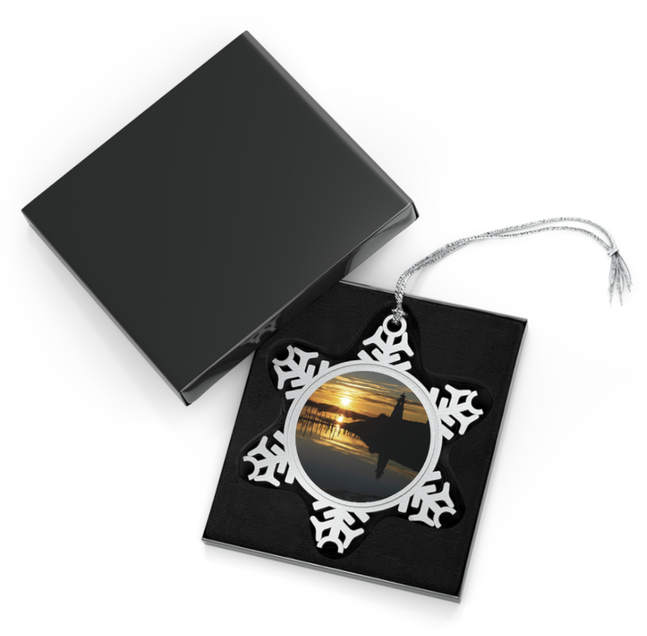 A snowflake-shaped pewter ornament featuring a photograph of the white Mulholland Point Lighthouse silhouetted against a vibrant sunrise, reflected in the calm water.