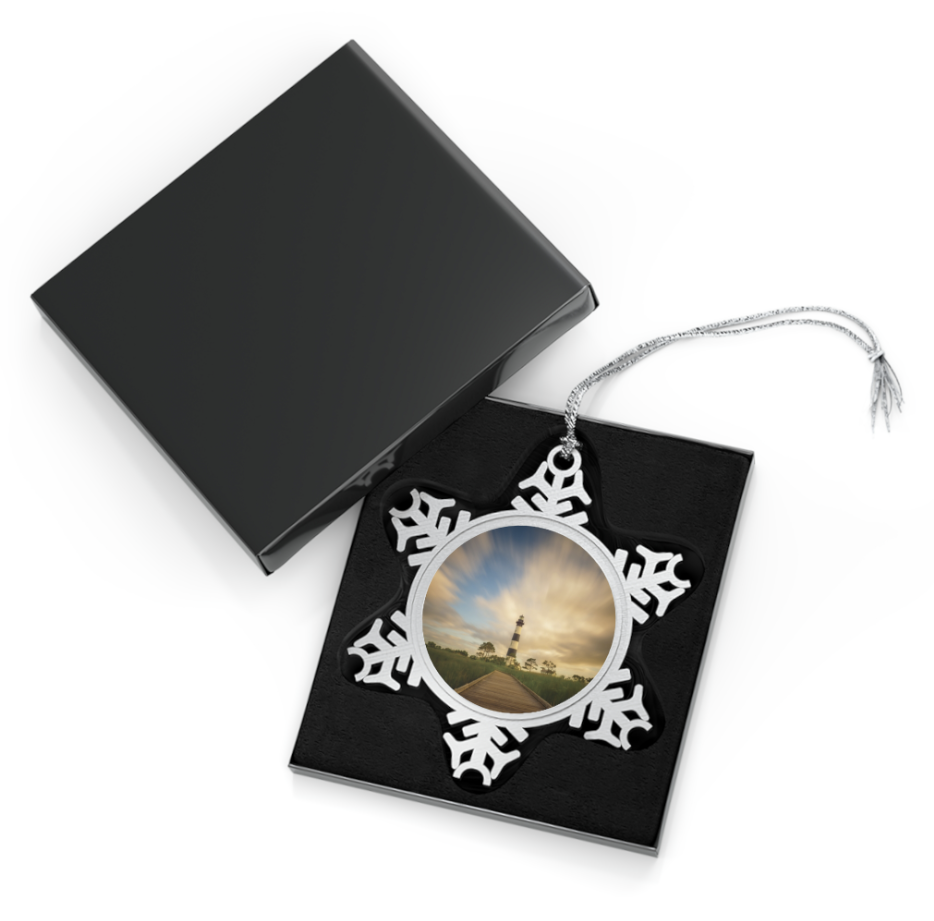 A snowflake-shaped pewter ornament featuring a photograph of the black and white striped Bodie Island Lighthouse, with long exposure clouds streaking across a partly blue, partly golden sky.