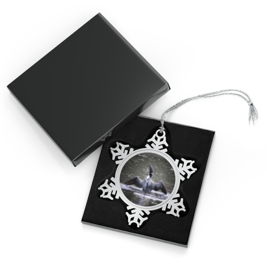 A snowflake-shaped pewter ornament featuring a photograph of a Common Loon rearing up in the water with its wings spread, creating a splash, showcasing its black and white patterned feathers.