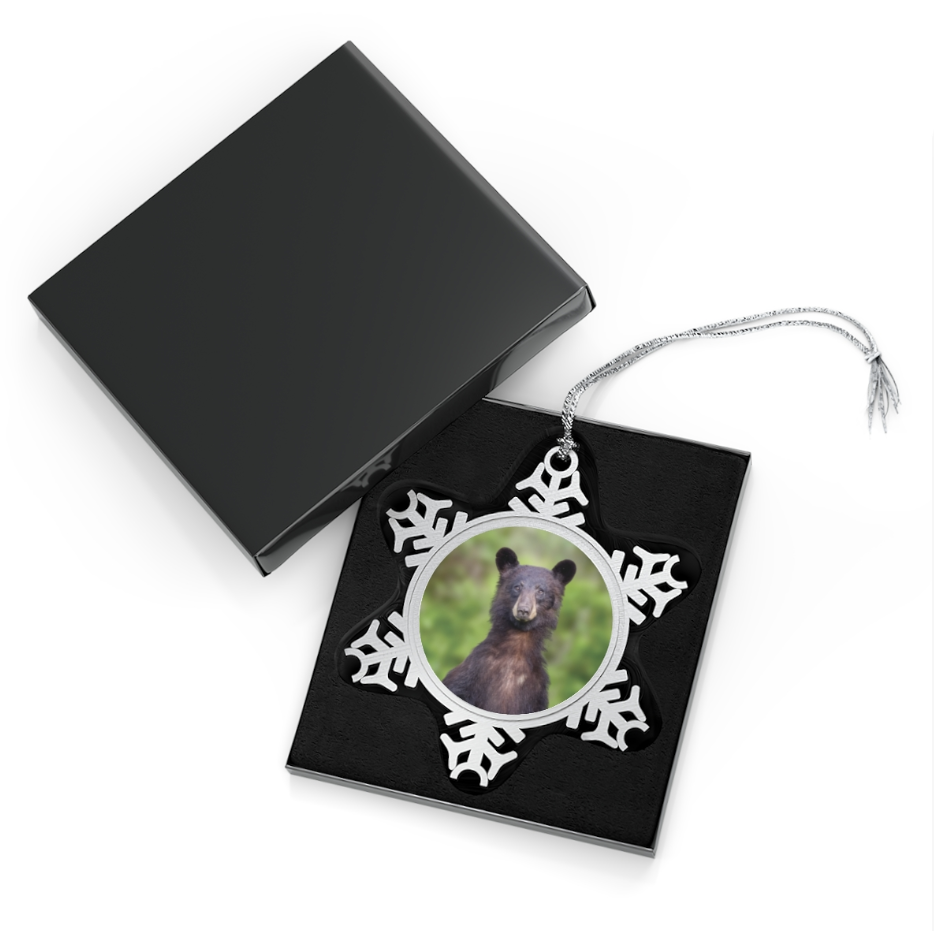 A snowflake-shaped pewter ornament featuring a photograph of a young black bear standing upright on its hind legs in a green, leafy natural environment, looking directly at the viewer.