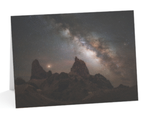 A stunning photo of the Milky Way galaxy over a desert landscape, with two rock formations resembling mule ears in the foreground. The night sky is filled with stars and nebulae, creating a contrast with the dark silhouette of the rocks and plants. The photo showcases the beauty and mystery of the cosmos, as well as the skill and creativity of Andy Morgan Photography.