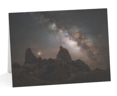 A stunning photo of the Milky Way galaxy over a desert landscape, with two rock formations resembling mule ears in the foreground. The night sky is filled with stars and nebulae, creating a contrast with the dark silhouette of the rocks and plants. The photo showcases the beauty and mystery of the cosmos, as well as the skill and creativity of Andy Morgan Photography.