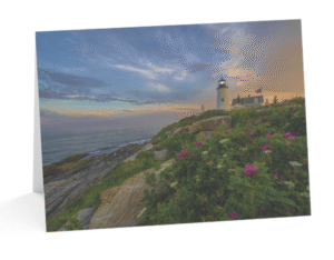 A notecard displaying a photograph of the white Pemaquid Point Lighthouse on a rocky cliff overlooking the ocean, surrounded by vibrant pink blossoms and green foliage under a colorful sunset sky.
