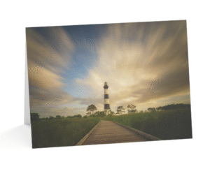 A notecard displaying a photograph of the black and white striped Bodie Island Lighthouse with long exposure clouds streaking across a partly blue, partly golden sky, and a wooden boardwalk leading towards it through tall grass.