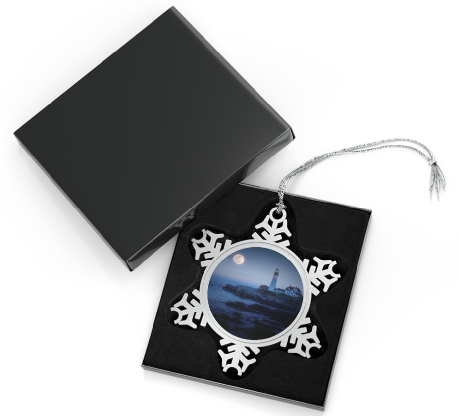 A snowflake-shaped pewter ornament featuring a photograph of Portland Head Light at night, with the moon visible in the sky.