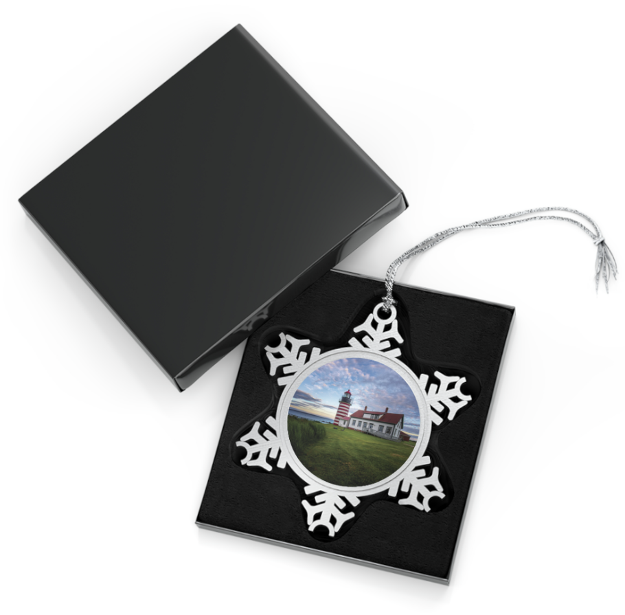 A snowflake-shaped pewter ornament featuring a photograph of the red and white striped West Quoddy Head Lighthouse, with a green lawn in front and a blue sky with clouds.