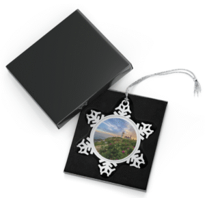 Silver snowflake ornament with a round photo insert featuring Pemaquid Point Lighthouse at sunset, overlooking the rocky coastline with wildflowers and green vegetation, displayed in an open black gift box with a black velvet interior.