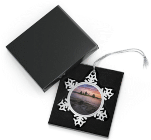 Silver snowflake ornament with a round photo insert featuring Pumpkin Island Lighthouse at sunset, seen across calm water with a rocky shoreline and silhouetted trees on the island, displayed in an open black gift box with a black velvet interior.