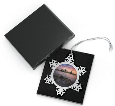 Silver snowflake ornament with a round photo insert featuring Pumpkin Island Lighthouse at sunset, seen across calm water with a rocky shoreline and silhouetted trees on the island, displayed in an open black gift box with a black velvet interior.