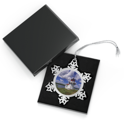 Silver snowflake ornament with a round photo insert featuring Squirrel Point Lighthouse on a sunny day, with white clouds in a blue sky, green grass in the foreground, and calm water behind, displayed in an open black gift box with a black velvet interior.