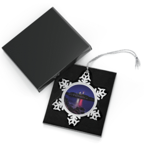 Silver snowflake ornament with a round photo insert featuring Cape Neddick Nubble Light at dusk or night, illuminated against a dark sky with reflections on the calm water and rocky foreground, displayed in an open black gift box with a black velvet interior.