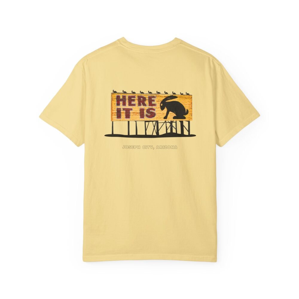 Light yellow t-shirt featuring a graphic of the "Here It Is" billboard sign from the Jackrabbit Trading Post on Route 66. The sign has a wooden texture with "HERE IT IS" in red letters and a large black silhouette of a jackrabbit. Smaller jackrabbit silhouettes are along the top. The text "JOSEPH CITY, ARIZONA" is written below the sign.