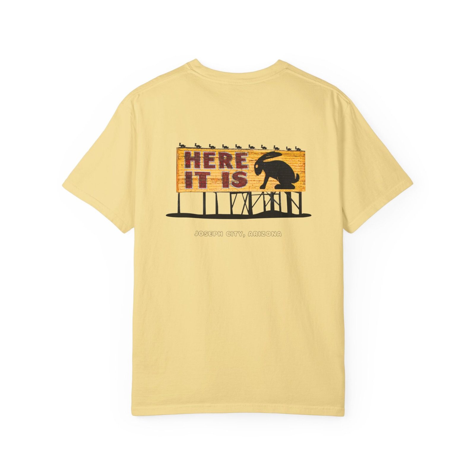 Light yellow t-shirt featuring a graphic of the "Here It Is" billboard sign from the Jackrabbit Trading Post on Route 66. The sign has a wooden texture with "HERE IT IS" in red letters and a large black silhouette of a jackrabbit. Smaller jackrabbit silhouettes are along the top. The text "JOSEPH CITY, ARIZONA" is written below the sign.