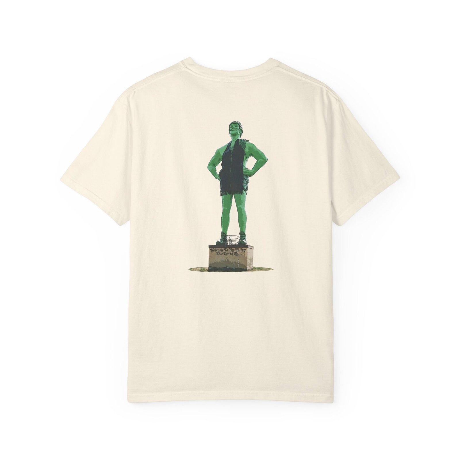 Cream-colored t-shirt featuring a graphic of the Jolly Green Giant statue in Blue Earth, Minnesota. The giant is green, wearing a dark green tunic, and standing on a pedestal. Text on the pedestal reads "WELCOME TO BLUE EARTH THE HOME OF THE JOLLY GREEN GIANT."