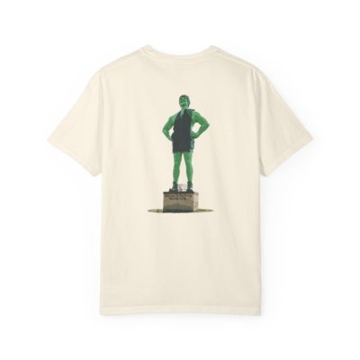 Cream-colored t-shirt featuring a graphic of the Jolly Green Giant statue in Blue Earth, Minnesota. The giant is green, wearing a dark green tunic, and standing on a pedestal. Text on the pedestal reads "WELCOME TO BLUE EARTH THE HOME OF THE JOLLY GREEN GIANT."