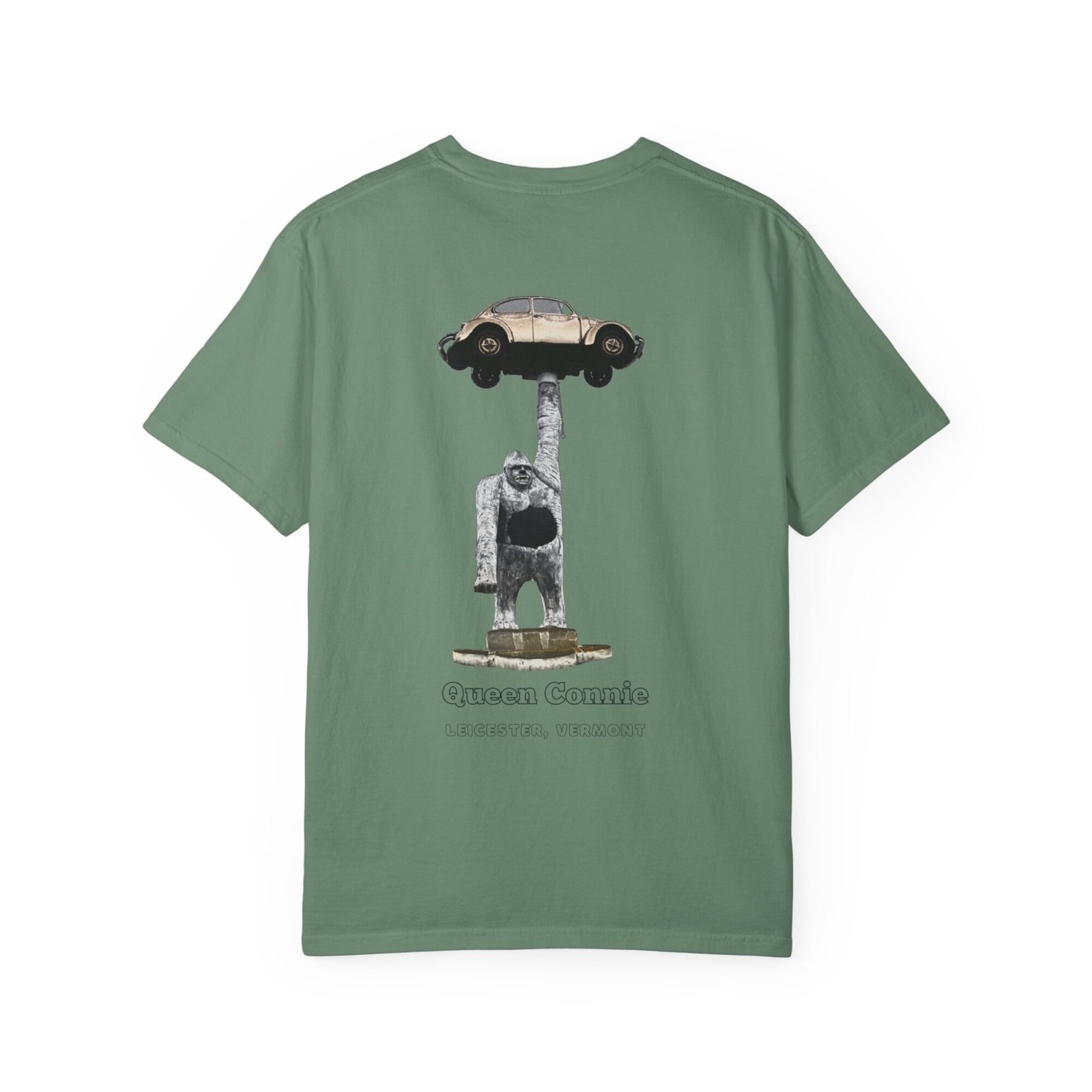 Olive green t-shirt featuring a graphic of the Queen Connie statue. The statue depicts a large white gorilla holding up a beige Volkswagen Beetle car above its head. Text below the graphic reads "Queen Connie" and "LEICESTER, VERMONT."
