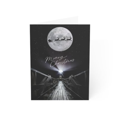 Christmas notecard featuring a high-contrast black-and-white photograph of Marshall Point Lighthouse and its wooden walkway under a full moon, with a silhouette of Santa's sleigh and the text "Merry Christmas" in elegant white script.