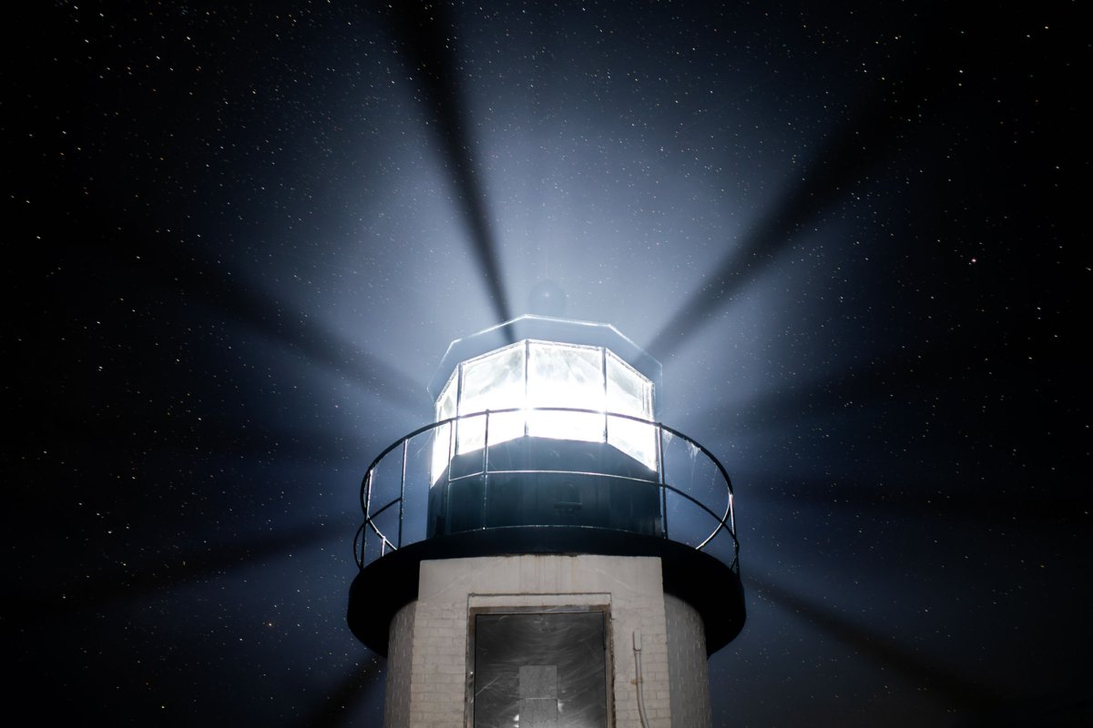 A close-up photograph looking up at the top of the Marshall Point Lighthouse in Port Clyde, Maine, with its powerful light beams fanning out against a clear, starry night sky.