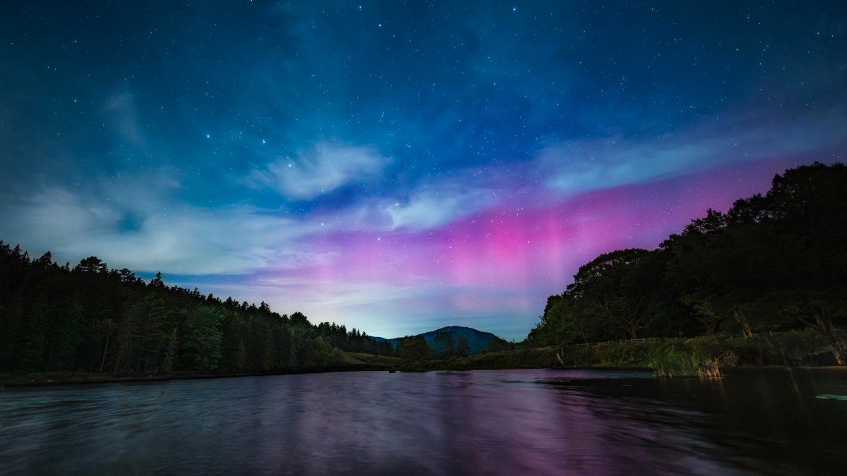 Long-exposure night photograph of the Northern Lights (Aurora Borealis) showing vibrant magenta and purple colors over a dark, still lake or river bordered by dense green forests in Acadia National Park.