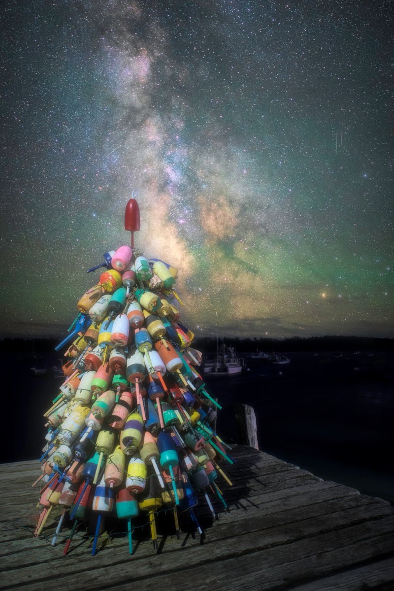 Night photograph of a brightly colored lobster buoy Christmas tree standing on a wooden dock in Friendship, Maine, with the luminous band of the Milky Way stretching across the dark sky above.