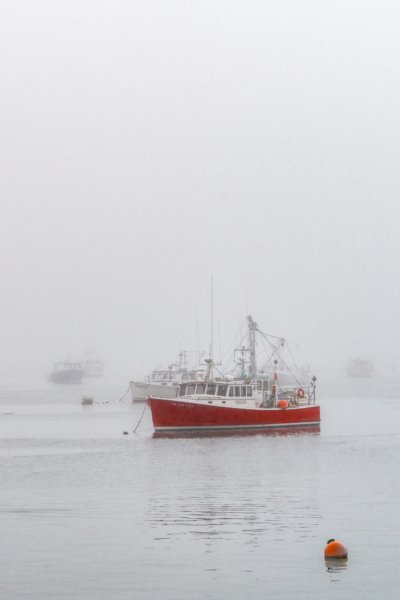 Fine art photograph of a bright red lobster boat moored in dense gray fog on the Maine coast, with several faint white boats visible in the background and an orange buoy in the foreground.